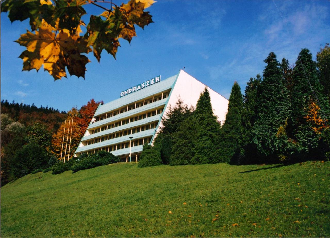 Piramidalny hotel 'Ondraszek' z niebieskim dachem, otoczony zielenią i drzewami w jesiennej scenerii pod błękitnym niebem. Liście klonu w górnym rogu kadru.