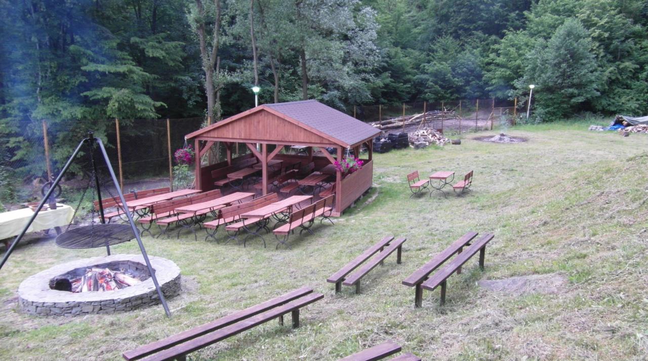 Plenerowa strefa rekreacyjna z drewnianą altaną, ławkami i paleniskiem na terenie zielonym. Grill zawieszony nad paleniskiem, w tle las.