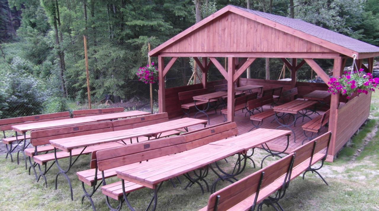 Zadaszona wiata z drewnianymi stołami i ławkami w plenerze, z kwiatami w donicach. Idealne miejsce na imprezy plenerowe, pikniki firmowe lub rodzinne spotkania na świeżym powietrzu.