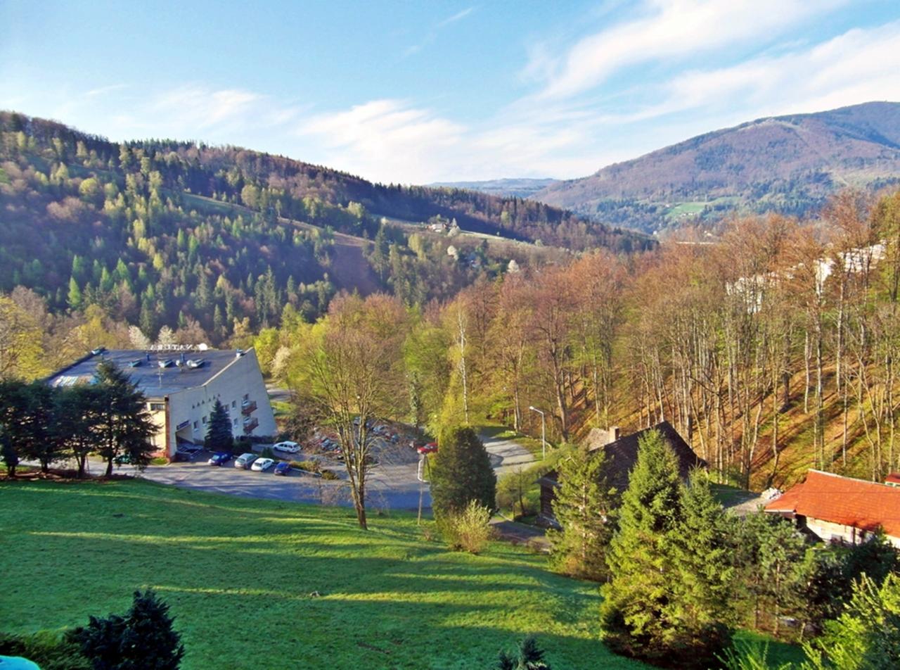 Krajobraz górski z budynkiem pensjonatu i parkingiem w otoczeniu zieleni i drzew w słoneczny dzień.