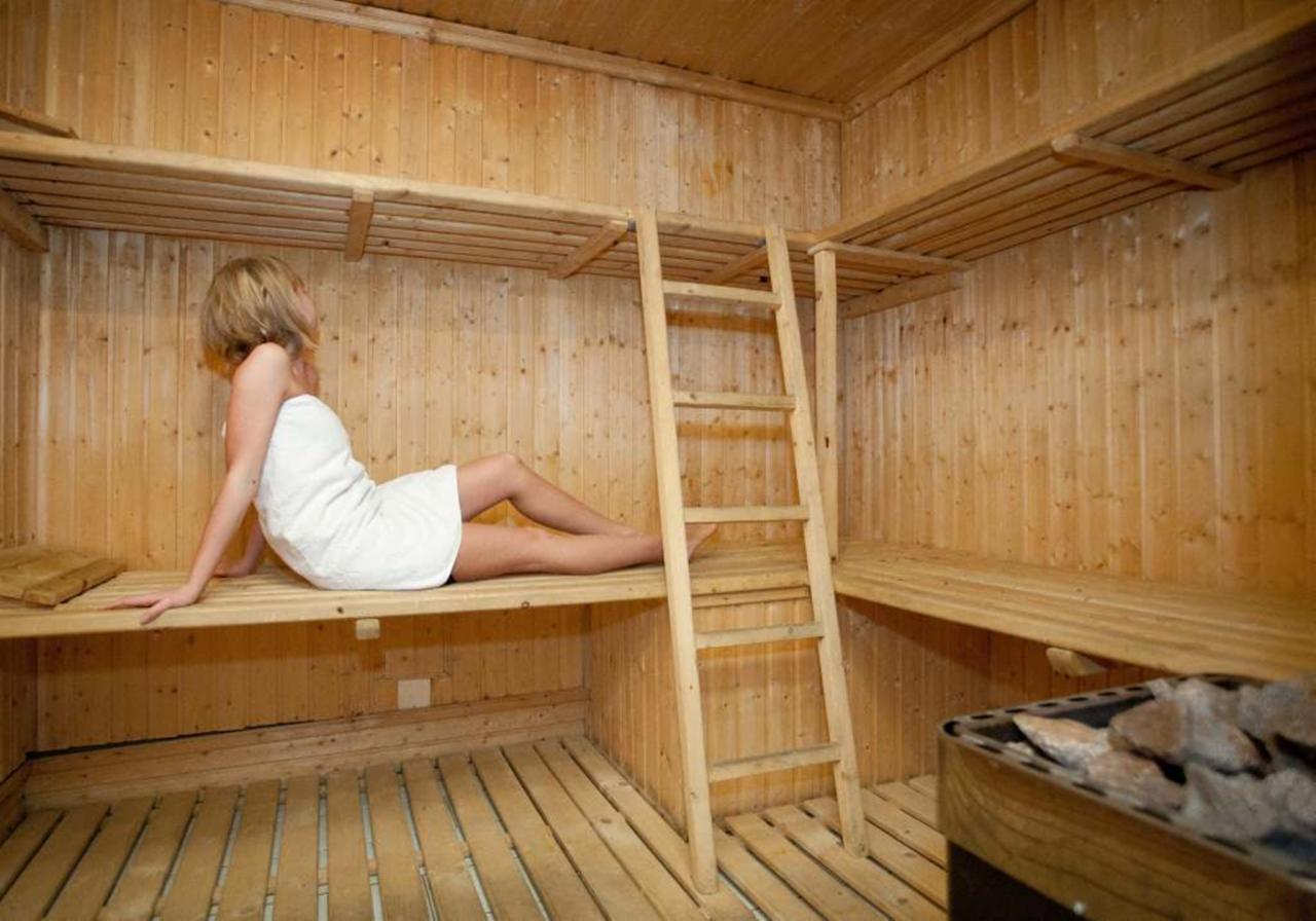 Kobieta w ręczniku relaksuje się w drewnianej saunie z drabinką i piecem z kamieniami. Ciepłe, stonowane światło podkreśla naturalny charakter wnętrza.
