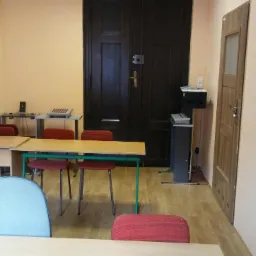 Gabinet z biurkami, krzesłami, i ciemnymi, dwuskrzydłowymi drzwiami w tle, sugerujący miejsce do terapii lub konsultacji.