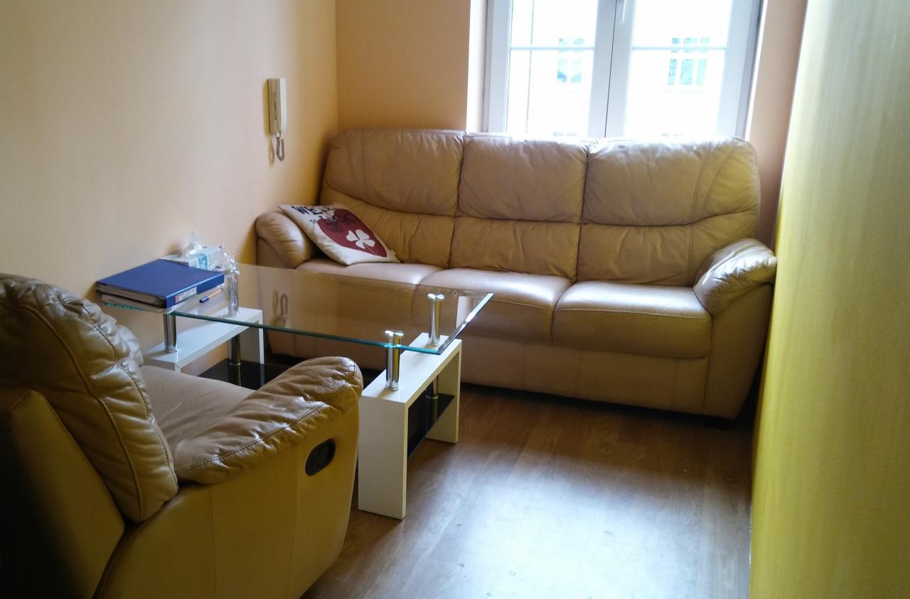 Jasny skórzany zestaw wypoczynkowy: sofa i fotel z funkcją relaks, stolik kawowy ze szklanym blatem, w tle okno i ściana z domofonem.