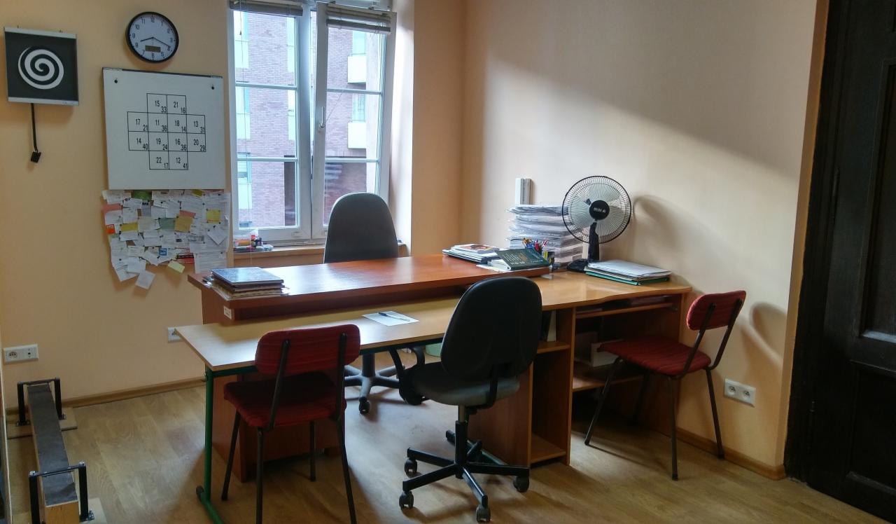 Gabinet z biurkiem, krzesłami, wentylatorem, tablicą z testami i notatkami na ścianie, zegarem i oknem z widokiem na budynek.