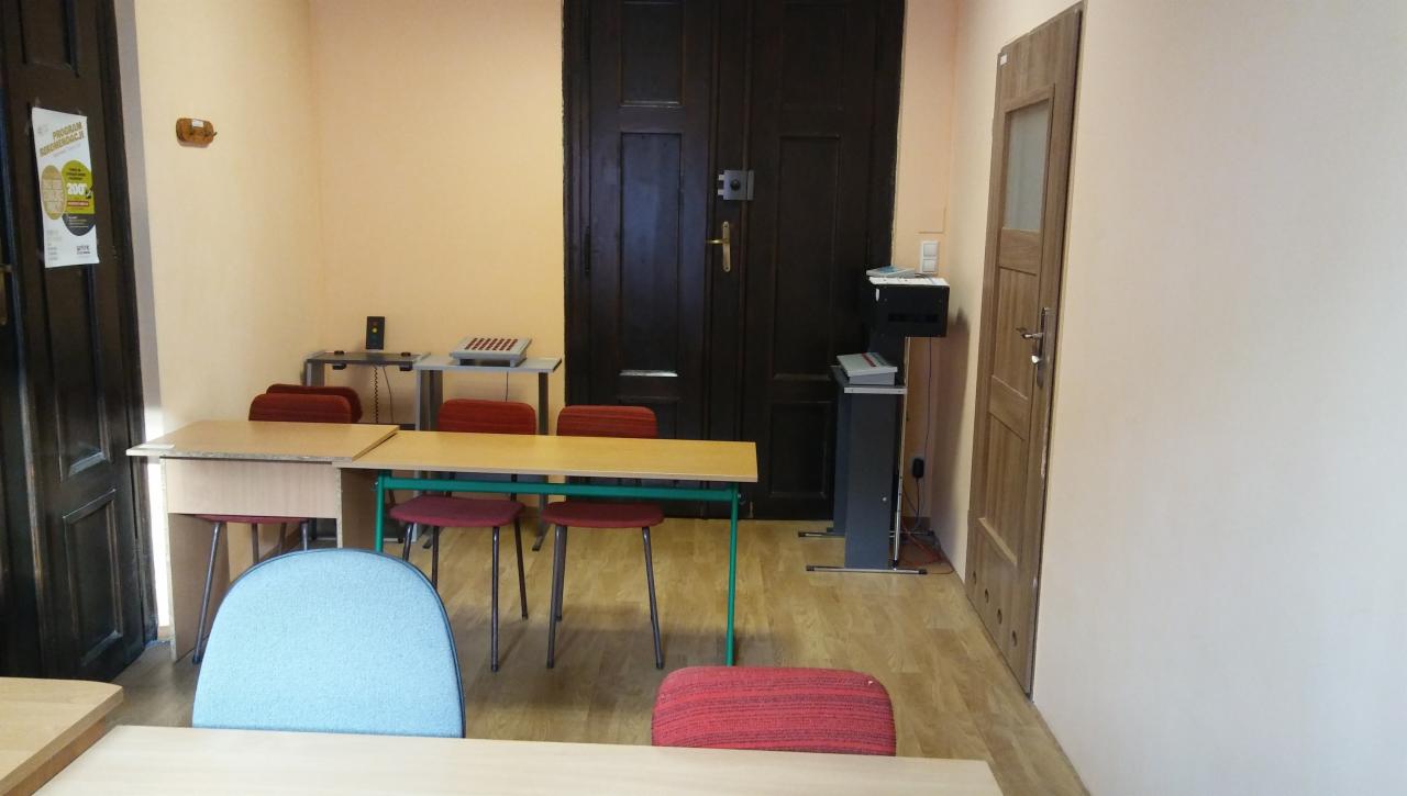 Gabinet z biurkami, krzesłami, i ciemnymi, dwuskrzydłowymi drzwiami w tle, sugerujący miejsce do terapii lub konsultacji.