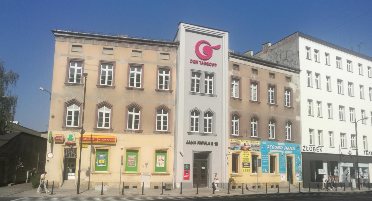 Fasada budynku Domu Targowego przy ulicy Jana Pawła II 18 z widocznym logo i szyldami różnych firm, w tym 'Badania Psychotechniczne'.