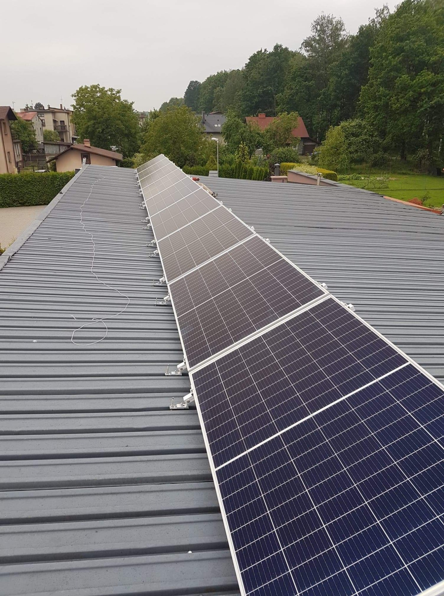 Rząd paneli słonecznych zamontowanych na szarym, metalowym dachu z widocznymi przewodami, w tle zabudowa jednorodzinna i drzewa w pochmurny dzień.