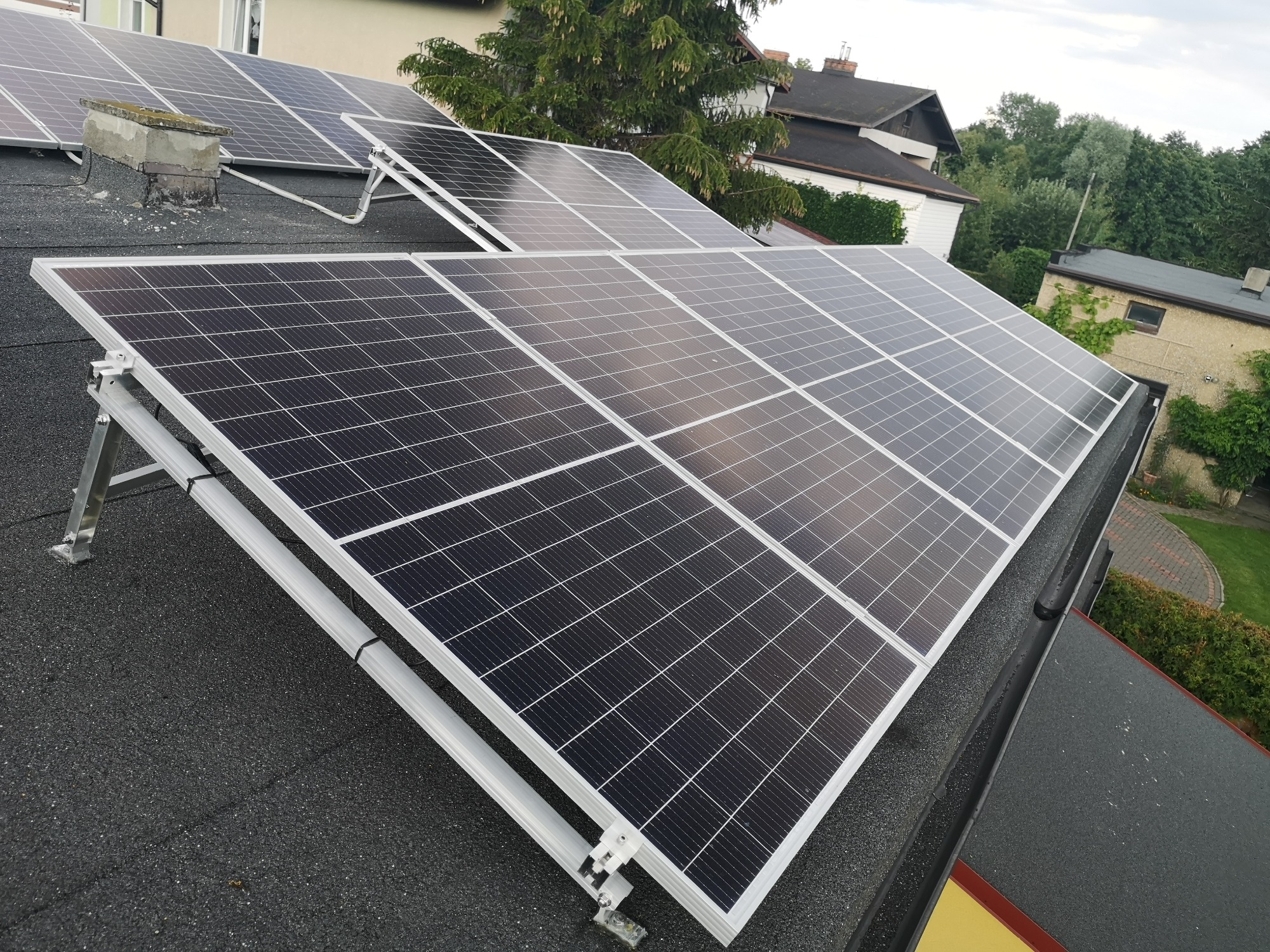 Panele słoneczne na dachu domu z kominem, zamontowane na aluminiowej konstrukcji, w tle zieleń drzew i zabudowa jednorodzinna.