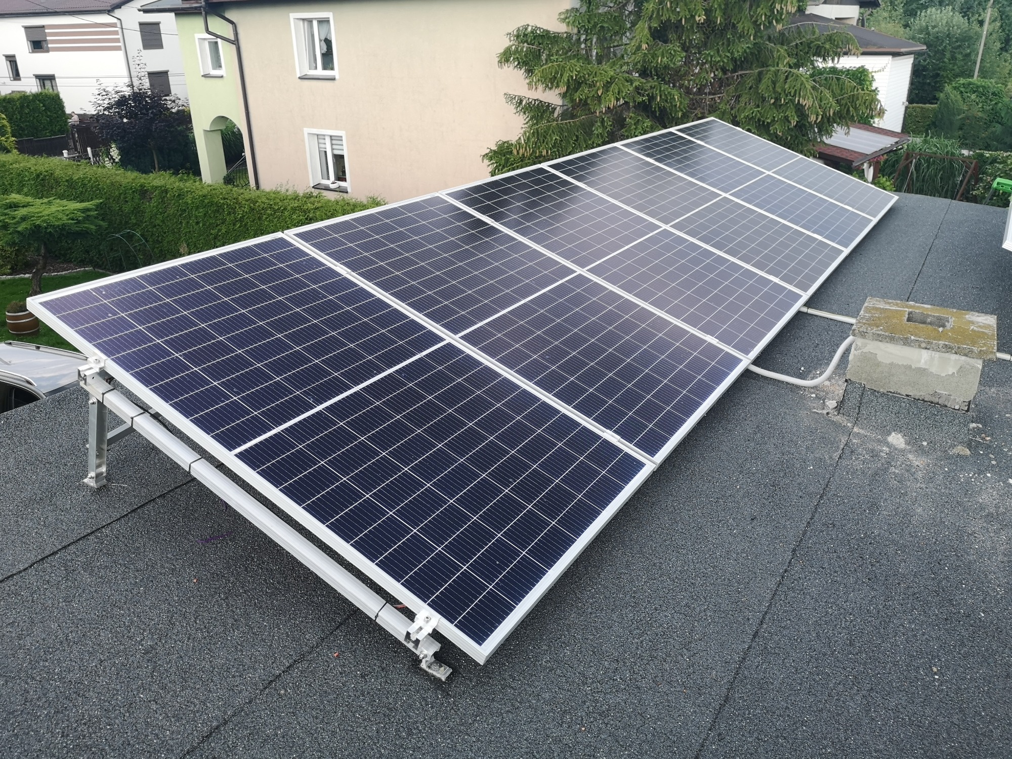 Panel fotowoltaiczny zamontowany na płaskim dachu pokrytym papą, widoczny z boku, na tle zieleni i zabudowań.