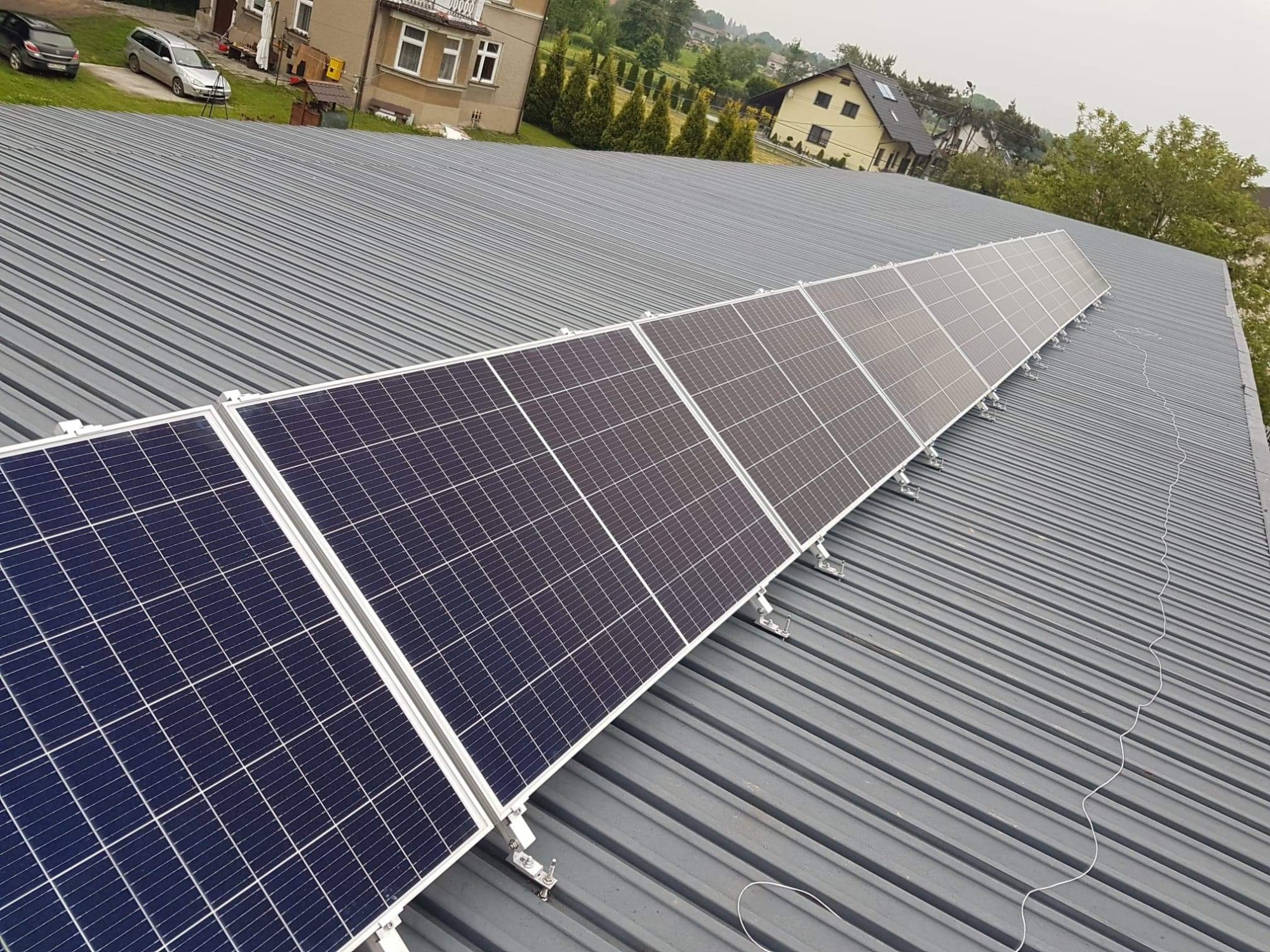 Rząd paneli fotowoltaicznych zamontowanych na szarym, metalowym dachu domu jednorodzinnego, widok z góry pod kątem.