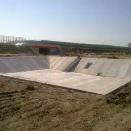 Betonowy zbiornik retencyjny w ziemi z zewnętrznymi schodami i otworami, widok z góry, w tle wiadukt. Jasne, naturalne światło.