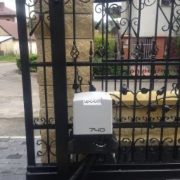 Czarna, ozdobna brama wjazdowa z automatycznym napędem FAAC 740, zamontowanym na słupku z piaskowca i kostce brukowej.