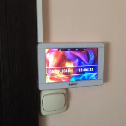 Panel sterowania alarmem Satel zamontowany na ścianie obok włącznika światła i futryny drzwi, wyświetlający datę i godzinę na tle abstrakcyjnej grafiki.