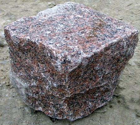 Czerwono-szary blok kostki granitowej o surowej fakturze, widoczny na tle piaszczystego podłoża, z wyraźnymi ziarnami minerałów.