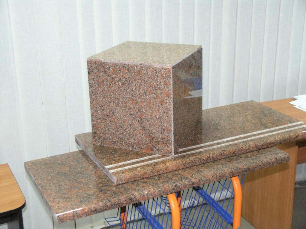 Polerowany blok z czerwono-brązowego granitu na blatach z tego samego materiału. Widoczny fragment biurka i zasłony w tle. Prezentacja próbki kostki granitowej.