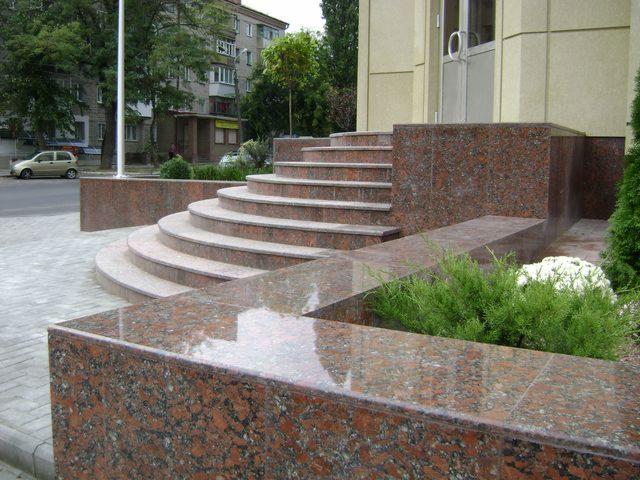 Wykończenie wejścia do budynku z czerwonego granitu: schody, murki i podjazd. Błyszcząca powierzchnia, staranne ułożenie, zieleń w tle. Estetyczne detale architektoniczne.