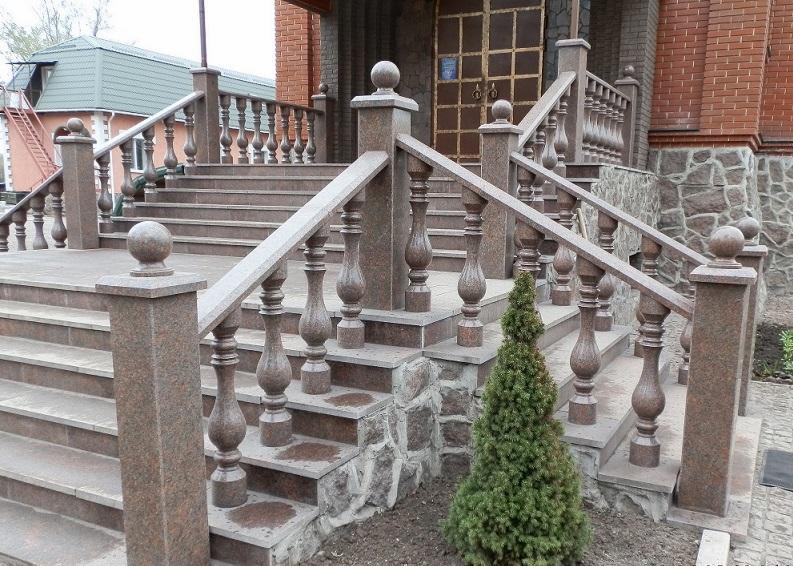 Granitowe schody wejściowe z balustradą i kulistymi zwieńczeniami, prowadzące do budynku z kamienną elewacją, uzupełnione małym drzewkiem iglastym.