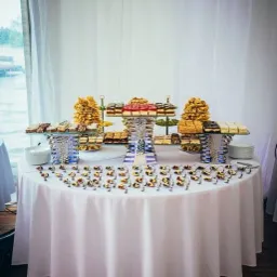 Stół z białym obrusem, na którym znajdują się różnorodne desery i ciasta na szklanych paterach o różnych wysokościach, a także małe babeczki z dekoracjami ułożone w rzędzie wzdłuż krawędzi stołu...