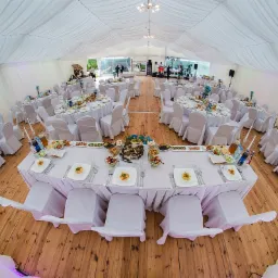 Uroczysta sala bankietowa w białym namiocie z drewnianą podłogą, nakryte stoły z białymi obrusami i krzesłami, na scenie zespół muzyczny.