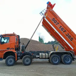 Pomarańczowa wywrotka KH-Kipper z podniesioną skrzynią ładunkową, na której widnieje numer telefonu 507 167 365, na tle hałdy żwiru i zakładu produkcyjnego z kominem.