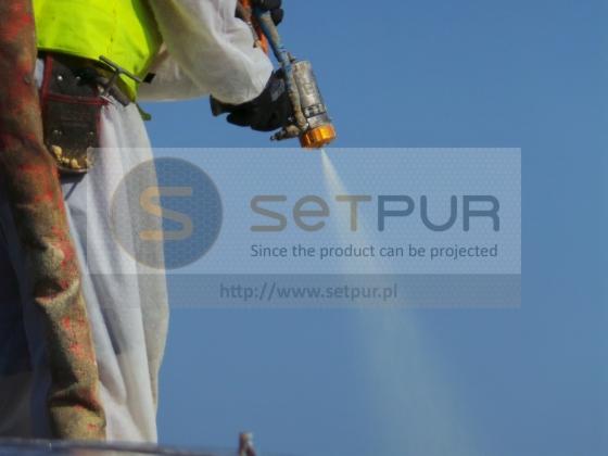 Pracownik w kombinezonie ochronnym natryskuje pianę PUR z pistoletu natryskowego na tle błękitnego nieba.