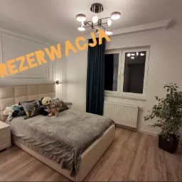 Nowoczesna sypialnia z beżowym łóżkiem, dekoracyjnymi poduszkami, pluszowymi misiami, ciemnozielonymi zasłonami i oryginalną lampą sufitową. Na ścianie widoczny napis 'REZERWACJA'.