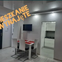 Jasne wnętrze mieszkania z widokiem na kuchnię, stół z czterema krzesłami, telewizor na ścianie, czarne drzwi.