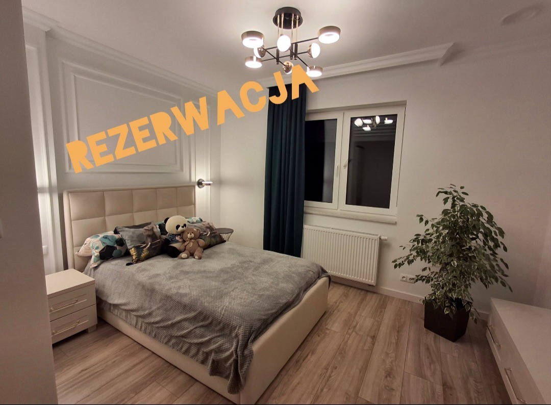 Nowoczesna sypialnia z beżowym łóżkiem, dekoracyjnymi poduszkami, pluszowymi misiami, ciemnozielonymi zasłonami i oryginalną lampą sufitową. Na ścianie widoczny napis 'REZERWACJA'.