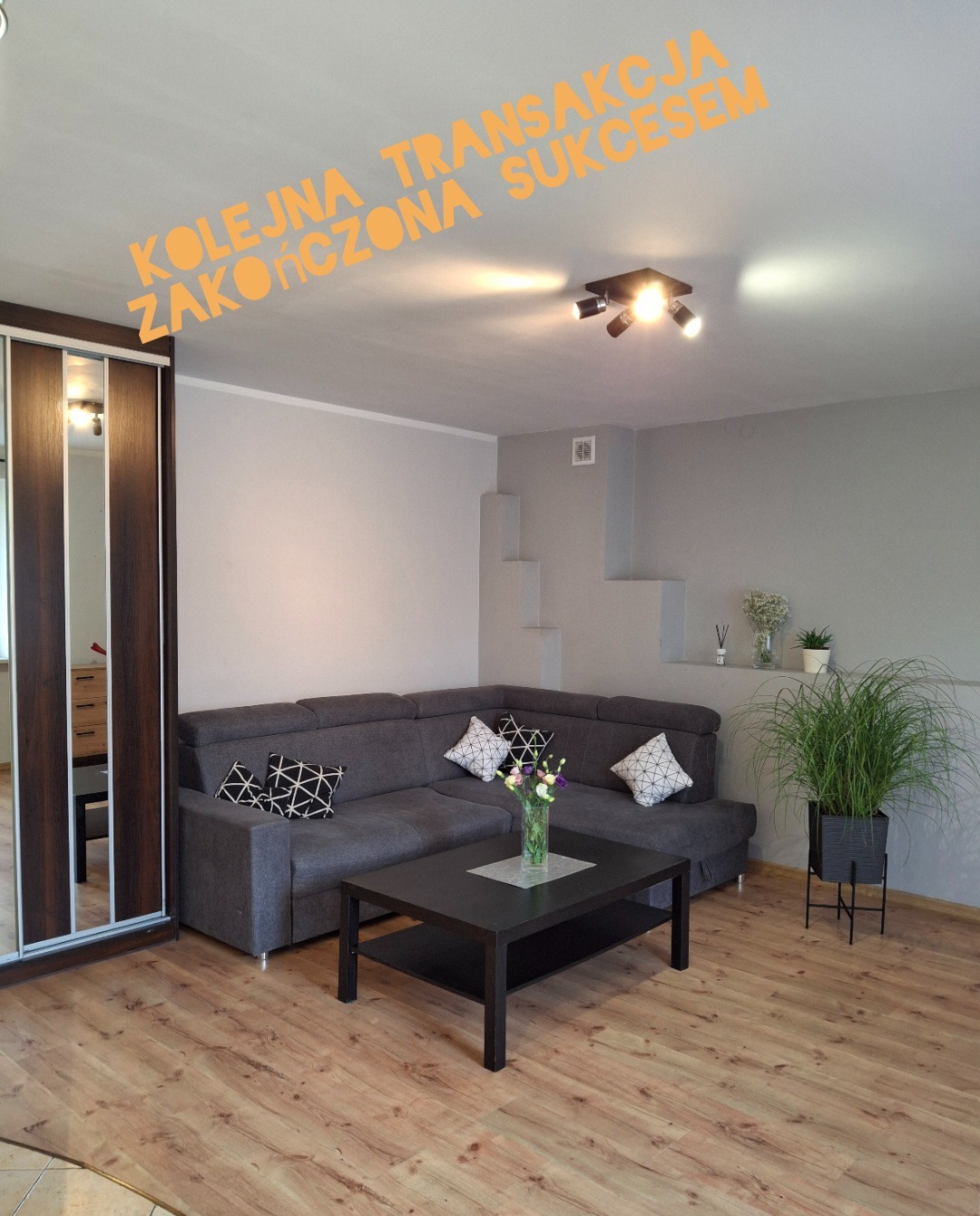 Umeblowany salon z narożną kanapą, stolikiem kawowym, przesuwną szafą i rośliną doniczkową, oświetlony trzema punktowymi lampami sufitowymi. Na zdjęciu widoczny napis 'Kolejna transakcja zakończona...