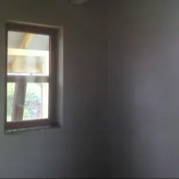 Świeżo nałożona, szara warstwa na ścianie wewnętrznej z widocznymi oknami z drewnianymi ramami, ukazująca częściowo konstrukcję dachu i zieleń na zewnątrz.