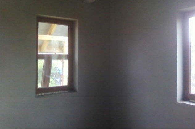 Świeżo nałożona, szara warstwa na ścianie wewnętrznej z widocznymi oknami z drewnianymi ramami, ukazująca częściowo konstrukcję dachu i zieleń na zewnątrz.