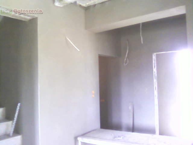 Świeżo otynkowane wnętrze z widocznymi przewodami elektrycznymi i fragmentem schodów w surowym stanie.