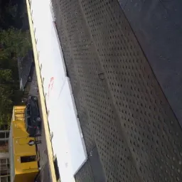 Dach z papy z perforacją i białą izolacją termiczną w trakcie montażu, widok z góry na budynek i samochód. Etap prac dekarskich.