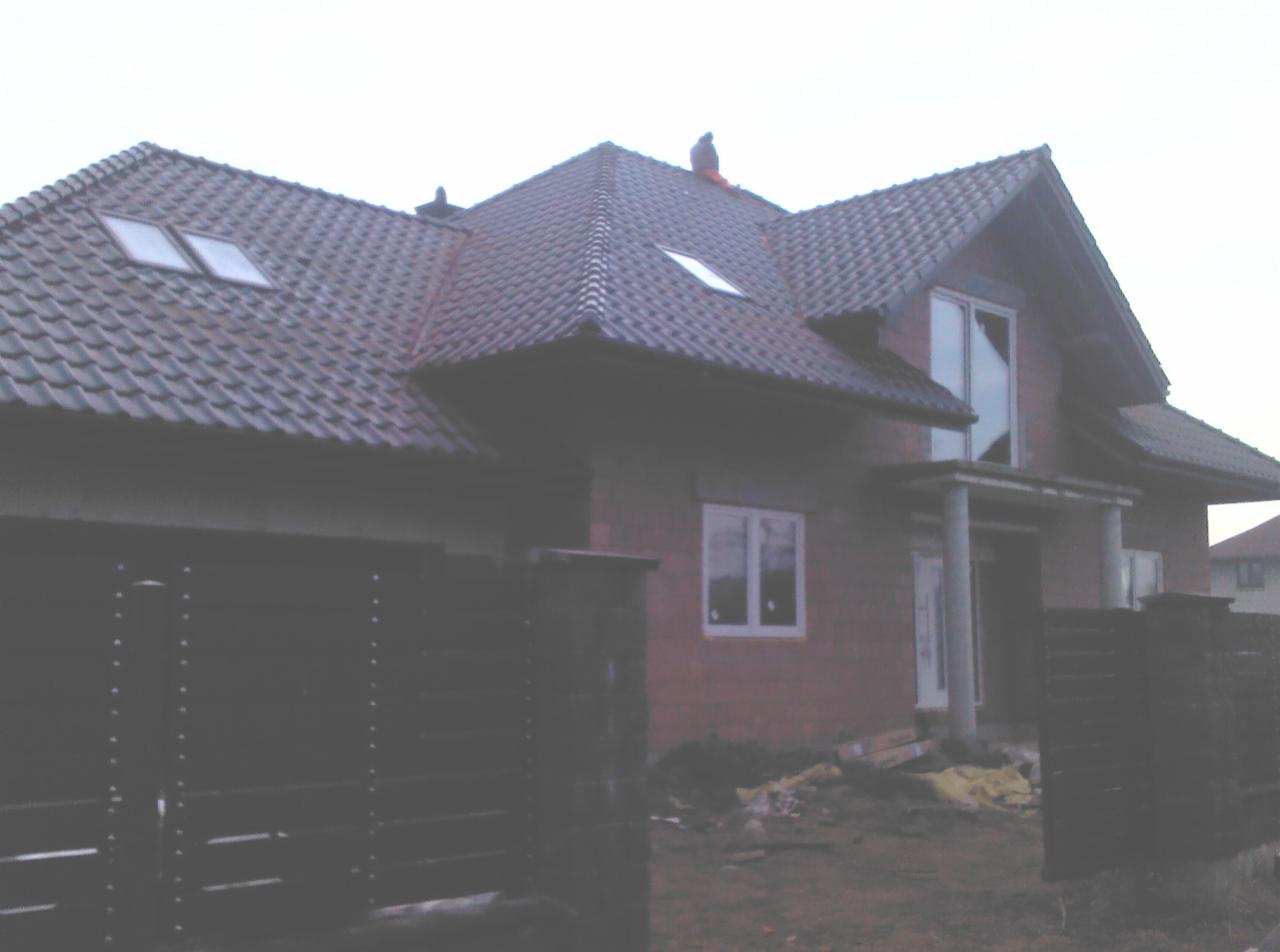 Dom w trakcie budowy z ciemną dachówką ceramiczną i oknami dachowymi. Widoczna brama wjazdowa i fragment elewacji z pustaków.