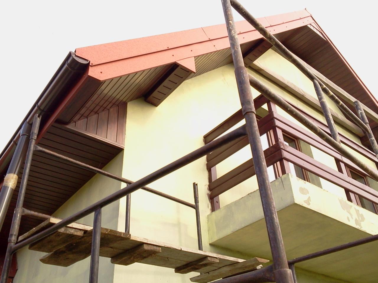 Dom z czerwonym dachem i brązowymi rynnami, widziany z rusztowania. Widoczny fragment elewacji i balkonu. Ujęcie z dołu podkreśla konstrukcję dachu.