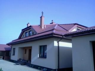 Dom z nowym, brązowym dachem wielospadowym i rynnami. Widoczne okna dachowe, komin i ozdobna figurka na szczycie. Jasna elewacja budynku.