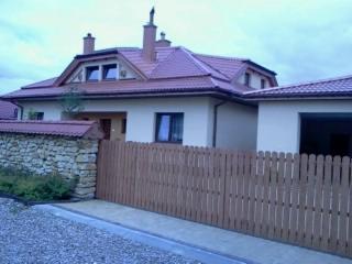 Dom z dachem z czerwonej dachówki, widoczny zza drewnianego płotu i kamiennego murku. Ujęcie z dołu, pochmurne niebo w tle.