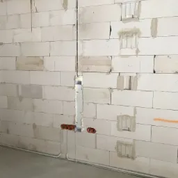 Wewnętrzna ściana z betonu komórkowego w trakcie prac elektrycznych: pionowy kanał kablowy wypełniony pianką montażową, widoczne pomarańczowe puszki instalacyjne i peszel przy podłodze.