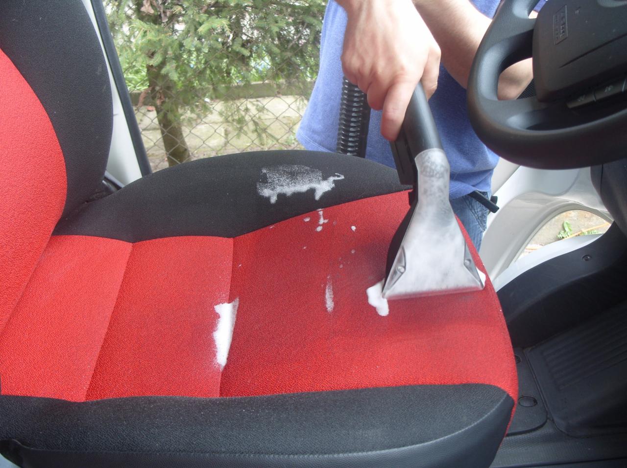 Czyszczenie tapicerki samochodowej odkurzaczem piorącym. Widoczne ślady piany na czerwono-czarnym fotelu. Ujęcie z wnętrza pojazdu, kierownica w tle.