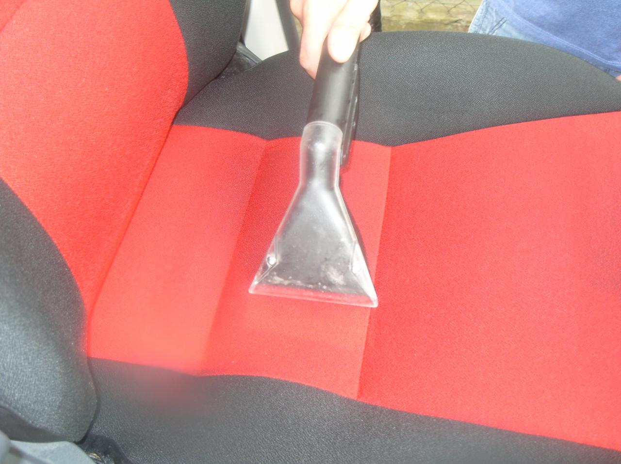 Czyszczenie tapicerki samochodowej na czerwono-czarnym fotelu za pomocą odkurzacza piorącego. Widoczna dysza i dłoń osoby wykonującej usługę.