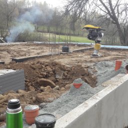 F.H.U. Ciapka Rafał Usługi Budowlane - Fundament domu w trakcie budowy: betonowe ściany, zbrojenie, ubijarka, rury kanalizacyjne. W tle drzewa i dym z ogniska.