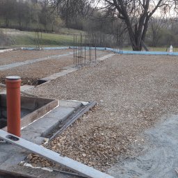 F.H.U. Ciapka Rafał Usługi Budowlane - Przygotowanie fundamentów pod dom: szalunek, podsypka żwirowa, pionowe rury kanalizacyjne. Widoczne zbrojenia i poziomica. Tło: zieleń wzgórz.