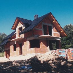 F.H.U. Ciapka Rafał Usługi Budowlane - Surowy stan domu z czerwonej cegły z czarnym dachem na tle błękitnego nieba i zieleni. Widoczne palety cegieł i mieszarka. Dom w trakcie budowy.