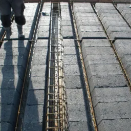 Betonowe bloczki stropowe zbrojone stalowymi prętami, widoczne buty pracownika w lewym górnym rogu.