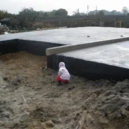 Świeżo wylana, betonowa płyta fundamentowa z izolacją pionową, na piaszczystym podłożu; mała dziewczynka w różowej kurtce przygląda się pracom.