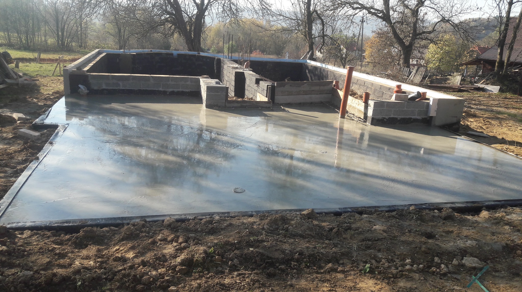 Wylany fundament pod budowę domu z widocznymi bloczkami betonowymi i wystającymi rurami kanalizacyjnymi. Świeży beton odbija światło słoneczne.