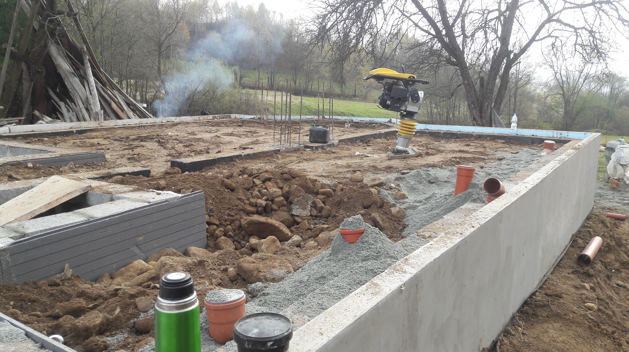 Fundament domu w trakcie budowy: betonowe ściany, zbrojenie, ubijarka, rury kanalizacyjne. W tle drzewa i dym z ogniska.