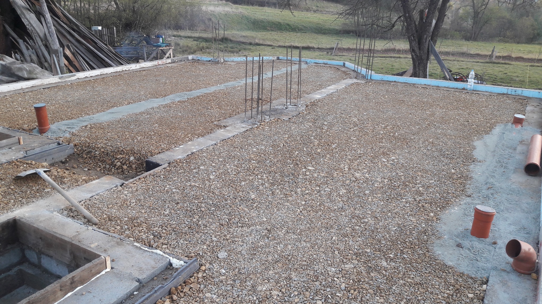 Fundament domu w trakcie budowy: podsypka żwirowa, zbrojenie, rury kanalizacyjne. Widoczne narzędzia i elementy szalunku. Tło: zielony teren.