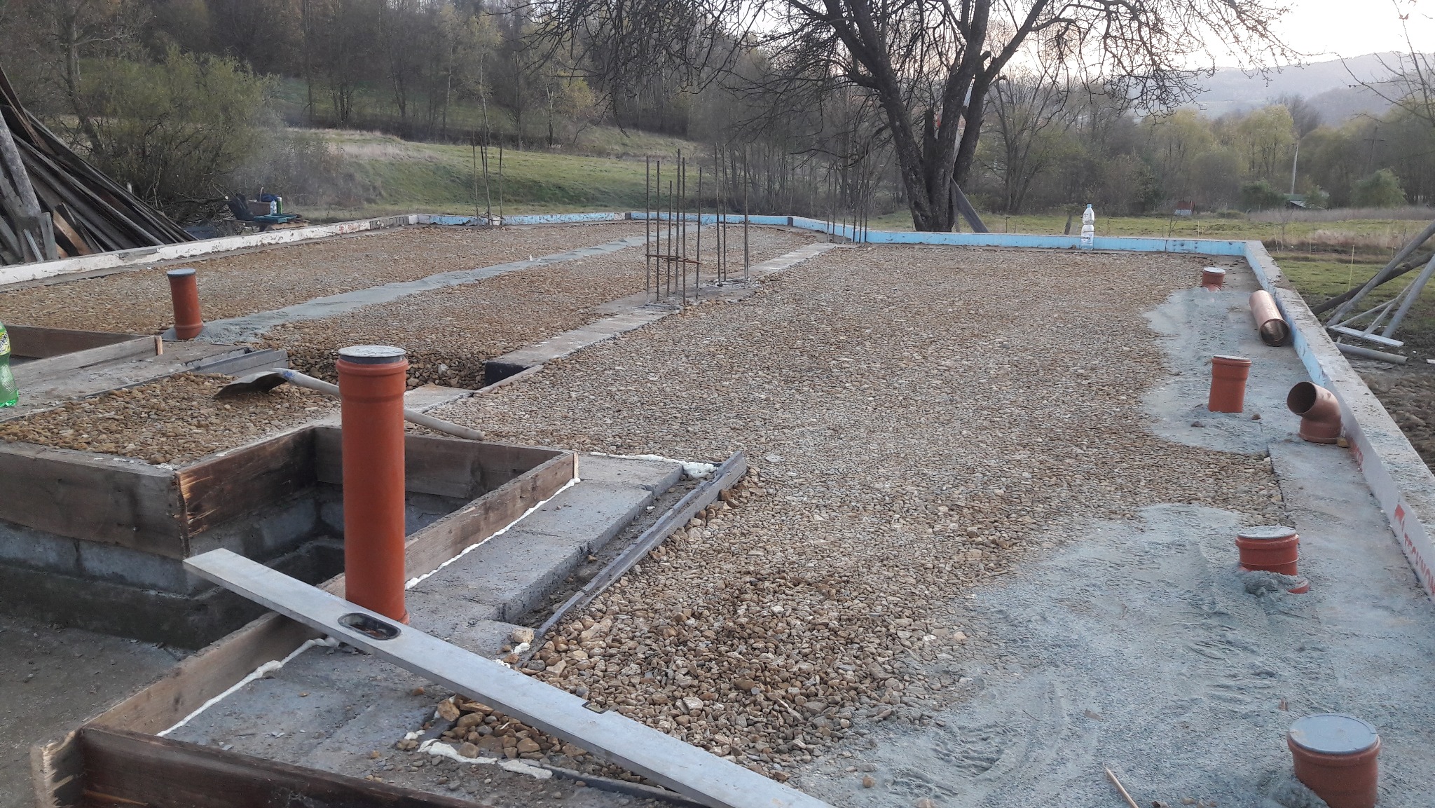 Przygotowanie fundamentów pod dom: szalunek, podsypka żwirowa, pionowe rury kanalizacyjne. Widoczne zbrojenia i poziomica. Tło: zieleń wzgórz.