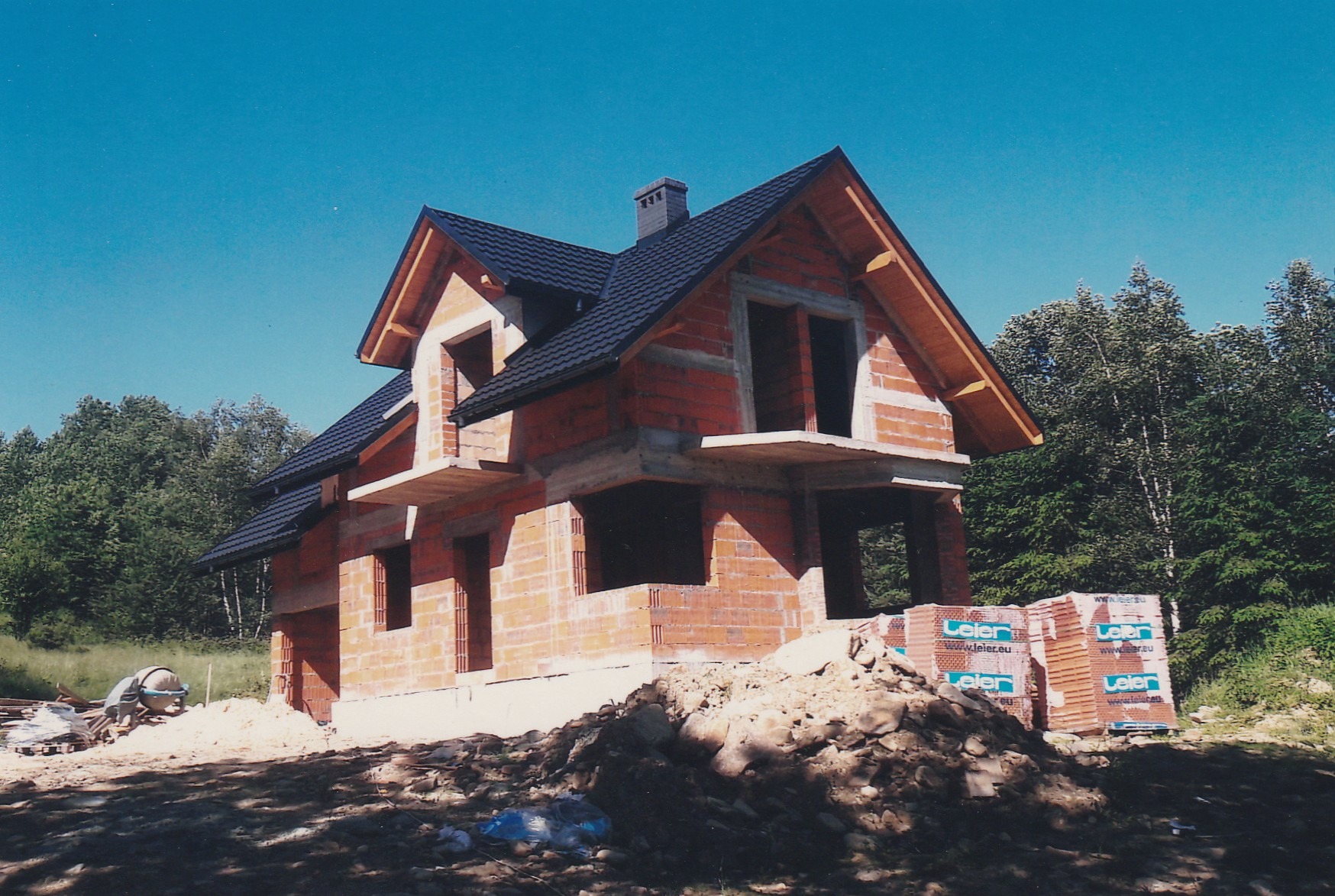 Surowy stan domu z czerwonej cegły z czarnym dachem na tle błękitnego nieba i zieleni. Widoczne palety cegieł i mieszarka. Dom w trakcie budowy.
